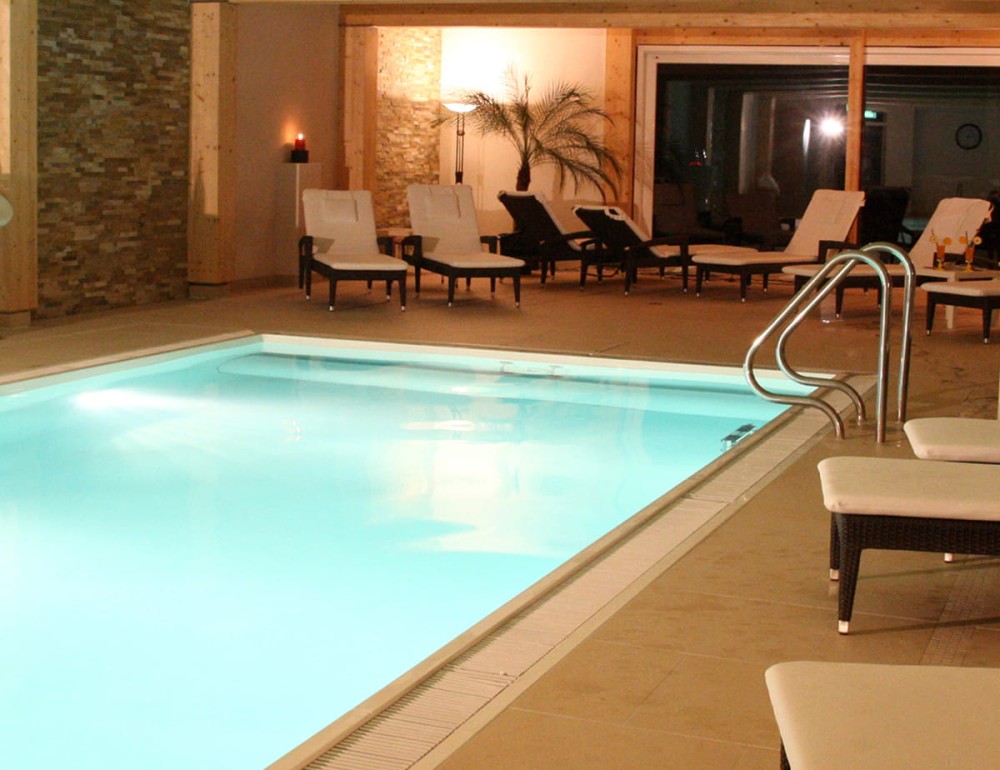 Beleuchtetes Hallenbad mit gem&uuml;tlichen Ruheliegen im Wellnessbereich