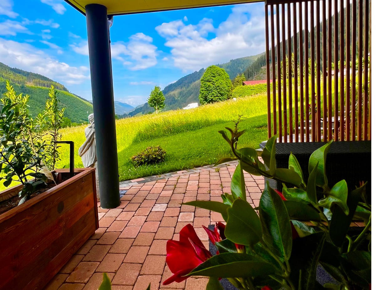 Terrasse mit Blick auf gr&uuml;ne Wiesen und bewaldete Berge unter blauem Himmel
