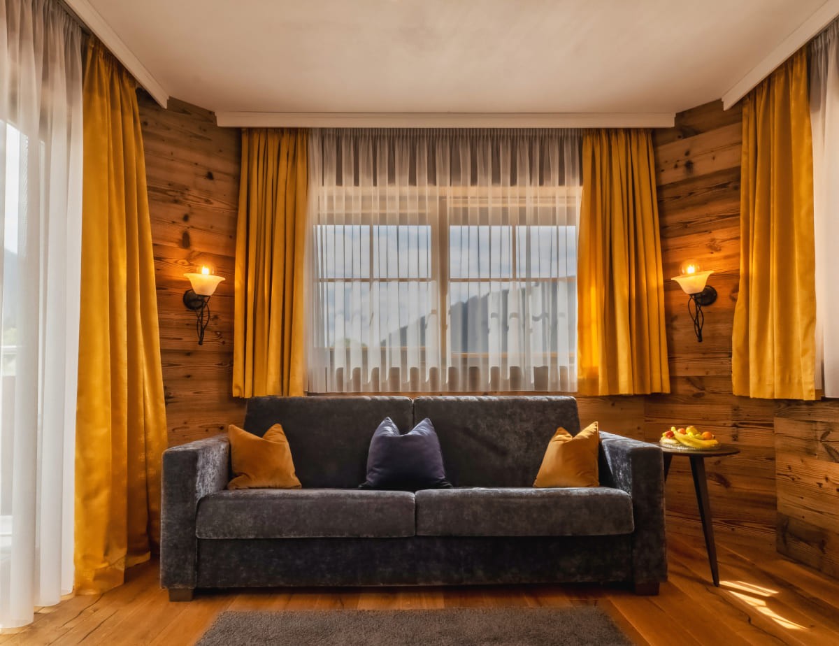Graues Sofa mit Kissen vor gelben Vorh&auml;ngen in einem gem&uuml;tlichen Raum mit Holzw&auml;nden