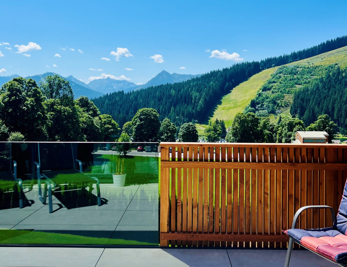 Blick von einer Terrasse auf bewaldete Berge und eine Wiese unter blauem Himmel